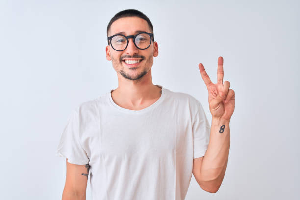 Young handsome man wearing glasses and standing over isolated background smiling with happy face winking at the camera doing victory sign. Number two. Young handsome man wearing glasses and standing over isolated background smiling with happy face winking at the camera doing victory sign. Number two. young man wink stock pictures, royalty-free photos & images