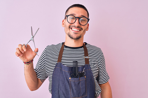 Young handsome hairdresser man wearing apron over pink isolated background with a happy face standing and smiling with a confident smile showing teeth