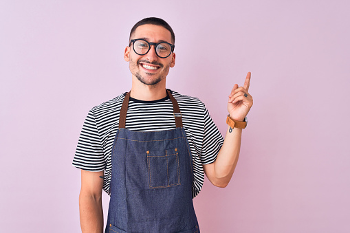 Young handsome man wearing employee apron over isolated background with a big smile on face, pointing with hand and finger to the side looking at the camera.