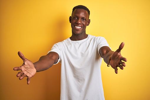 Young african american man wearing white t-shirt standing over isolated yellow background looking at the camera smiling with open arms for hug. Cheerful expression embracing happiness.