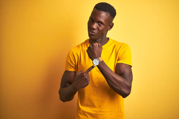 young african american man wearing casual t-shirt standing over isolated yellow background in hurry pointing to watch time, impatience, looking at the camera with relaxed expression - clock face clock deadline human hand imagens e fotografias de stock