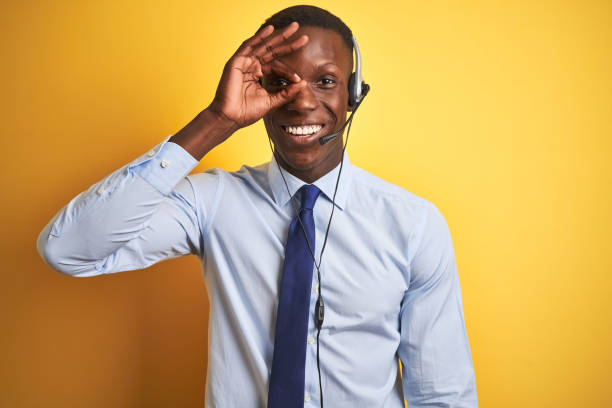 istock African american operator man working using headset over isolated yellow background doing ok gesture with hand smiling, eye looking through fingers with happy face. 1185936827