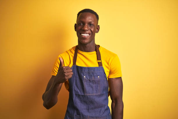 istock African american bartender man wearing apron standing over isolated yellow background doing happy thumbs up gesture with hand. Approving expression looking at the camera with showing success. 1185938898