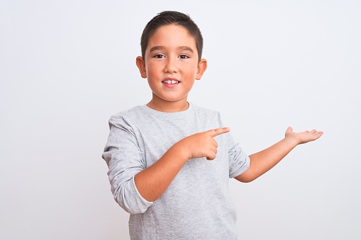 Beautiful kid boy wearing grey casual t-shirt standing over isolated white background amazed and smiling to the camera while presenting with hand and pointing with finger.