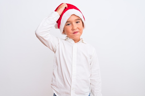Beautiful kid boy wearing Christmas Santa hat standing over isolated white background confuse and wonder about question. Uncertain with doubt, thinking with hand on head. Pensive concept.