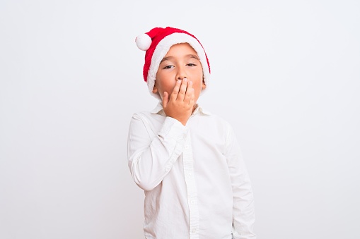 Beautiful kid boy wearing Christmas Santa hat standing over isolated white background bored yawning tired covering mouth with hand. Restless and sleepiness.