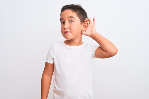 Beautiful kid boy wearing casual t-shirt standing over isolated white background smiling with hand over ear listening an hearing to rumor or gossip. Deafness concept.