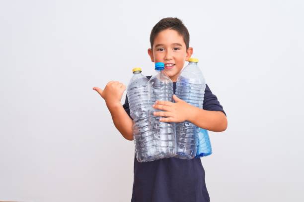 Beautiful kid boy recycling plastic bottles standing over isolated white background pointing and showing with thumb up to the side with happy face smiling