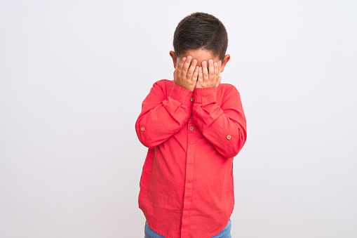 Beautiful kid boy wearing elegant red shirt standing over isolated white background with sad expression covering face with hands while crying. Depression concept.