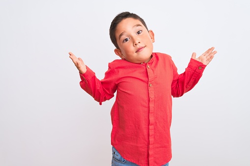 Beautiful kid boy wearing elegant red shirt standing over isolated white background clueless and confused expression with arms and hands raised. Doubt concept.