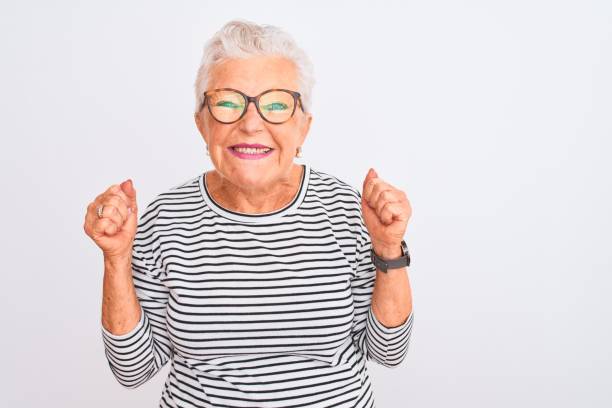 anziana donna dai capelli grigi che indossa occhiali da t-shirt navy a righe su sfondo bianco isolato eccitata per il successo con le braccia alzate e gli occhi chiusi che celebrano la vittoria sorridendo. concetto di vincitore. - women standing fist success foto e immagini stock