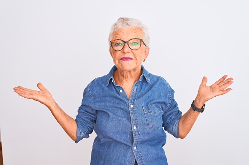Senior grey-haired woman wearing denim shirt and glasses over isolated white background smiling showing both hands open palms, presenting and advertising comparison and balance