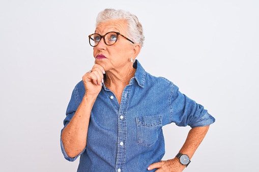 Senior grey-haired woman wearing denim shirt and glasses over isolated white background with hand on chin thinking about question, pensive expression. Smiling with thoughtful face. Doubt concept.