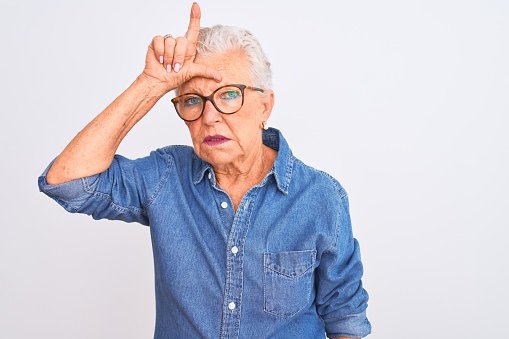 Senior grey-haired woman wearing denim shirt and glasses over isolated white background making fun of people with fingers on forehead doing loser gesture mocking and insulting.