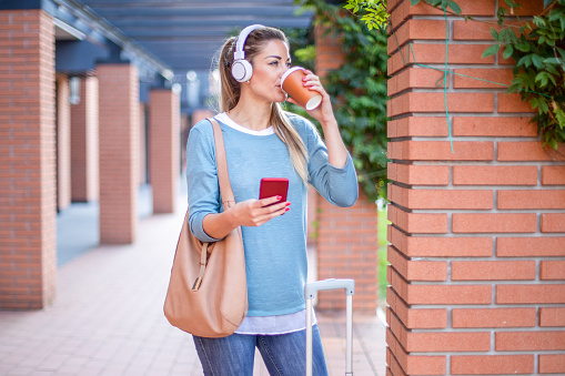 Portrait of beautiful young woman listening to music, drinking coffee and holding cell phone  in the street