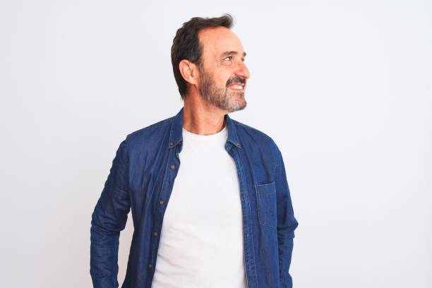 l'homme beau d'âge moyen utilisant la chemise bleue de denim restant au-dessus du sourire blanc d'isolement de fond regardant au côté et regardant loin pensant. - mannered photos et images de collection