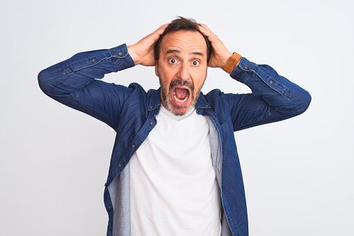 Middle age handsome man wearing blue denim shirt standing over isolated white background Crazy and scared with hands on head, afraid and surprised of shock with open mouth