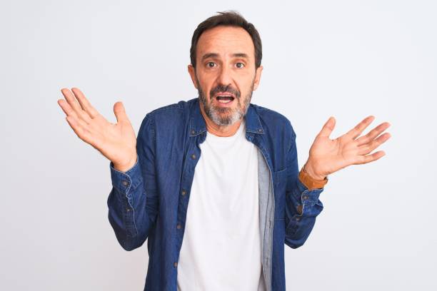 istock Middle age handsome man wearing blue denim shirt standing over isolated white background clueless and confused expression with arms and hands raised. Doubt concept. 1185950422