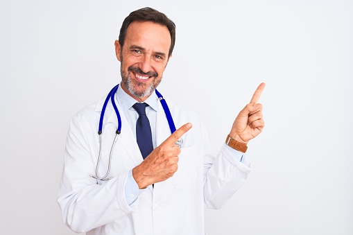 Middle age doctor man wearing coat and stethoscope standing over isolated white background smiling and looking at the camera pointing with two hands and fingers to the side.