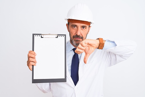 Senior engineer man wearing security helmet holding clipboard over isolated white background with angry face, negative sign showing dislike with thumbs down, rejection concept