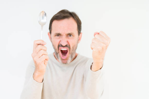 старший человек, держащий серебряную ложку над изолированным фоном раздраженный и разочарованный крик с гневом, сумасшедший и кричать с по - silver spoon in mouth стоковые фото и изображения