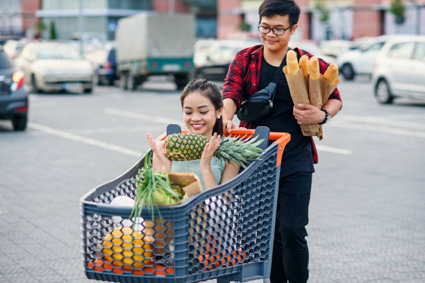 Cute asian girl holds pineapple and sits inside the shopping tray with food while vietnamese boy pushing it to their car on the parking near shopping centre. Cute happy asian girl holds pineapple and sits inside the shopping tray with food while vietnamese boy pushing it to their car on the parking near shopping centre. vietnamese girls for sale stock pictures, royalty-free photos & images