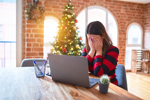 Beautiful woman sitting at the table working with laptop at home around christmas tree with sad expression covering face with hands while crying. Depression concept.