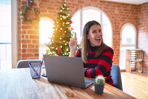 Beautiful woman sitting at the table working with laptop at home around christmas tree pointing finger up with successful idea. Exited and happy. Number one.