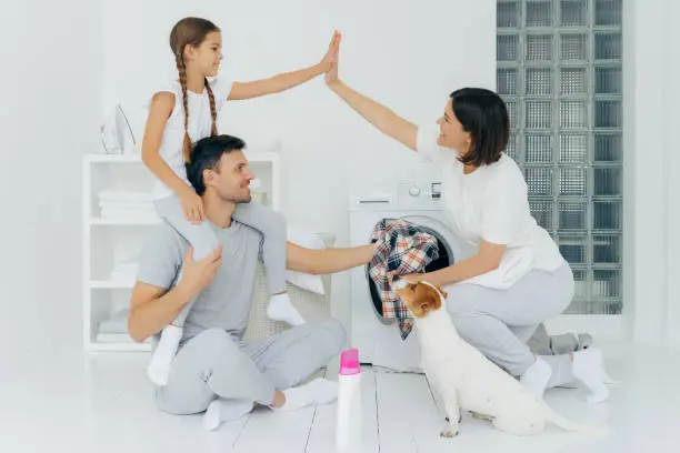 Photo of Well done! Daughter and mother make high five, rejoice almost finishing domestic work, girl rides on dads shoulders, wash clothing in laundry room with white walls. Jack russell terrier looks at host