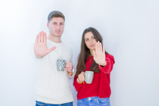 Beautiful young couple over white isolated background drinking a cup of coffee with open hand doing stop sign with serious and confident expression, defense gesture