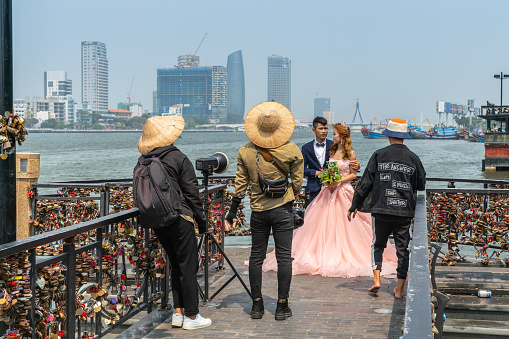 Da Nang, Vietnam - March 10, 2019: Bridal photo shoot on Love Bridge at Han River with downtown skyscrapers and fishing vessels in back. Photography crew, groom and bride in peach gown.