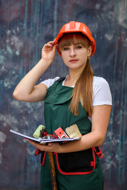 Woman in coverall and helmet holding notepad and posing for camera on grey background