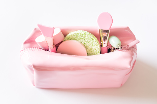 Pink cosmetic bag full of beauty products, silicon brushes, jade roller and different shapes sponges on white background. Skin care concept.