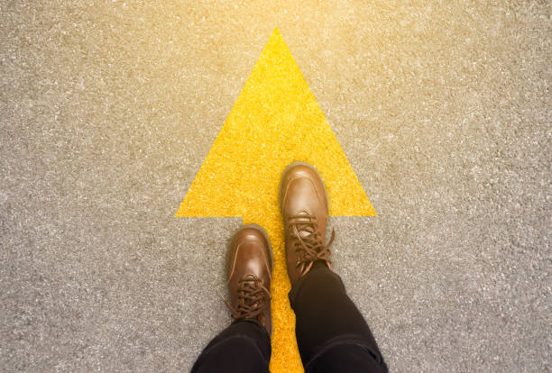 pieds et flèches sur le fond de route dans l'idée de commencement de ligne de départ. vue du haut. femme dans les bottines en cuir sur la voie avec le symbole jaune de flèche de direction. aller de l'avant, nouveau concept de départ et de réussite. - down view photos et images de collection