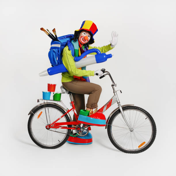 Female clown with backpack on his back, with paints and brushes, rides bicycle. A painted face, hat and big shoes. Studio shot.