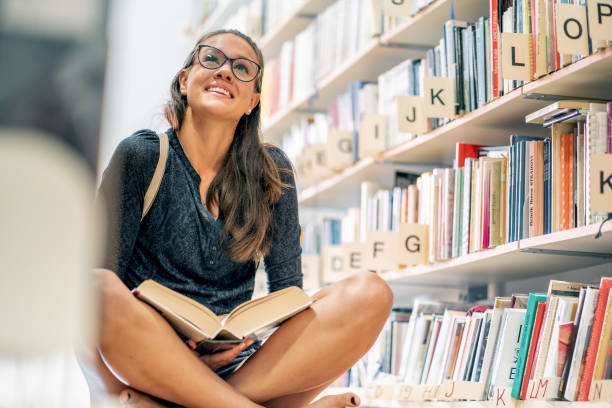 istock Smiling Young Woman Sitting on Floor in Library and Reading a Book 1186041704