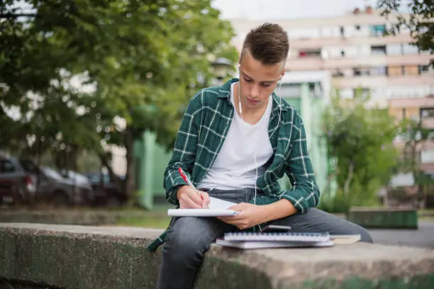 Teenager writing in his notebook and listening music with in-ear headphones in the schoolyard.