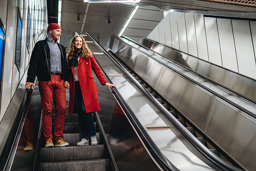 Young well dressed heterosexual couple holding hands while standing on escalator at subway station.