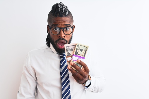 African american businessman with braids holding dollars over isolated white background scared in shock with a surprise face, afraid and excited with fear expression