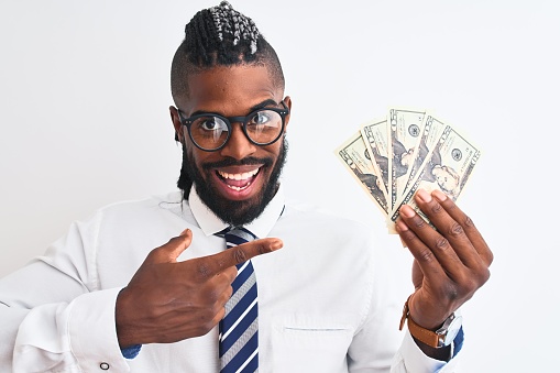 African american businessman with braids holding dollars over isolated white background very happy pointing with hand and finger
