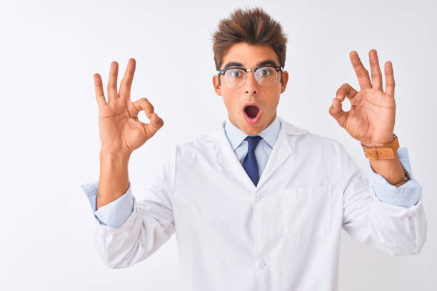 istock Young handsome sciencist man wearing glasses and coat over isolated white background looking surprised and shocked doing ok approval symbol with fingers. Crazy expression 1186061353