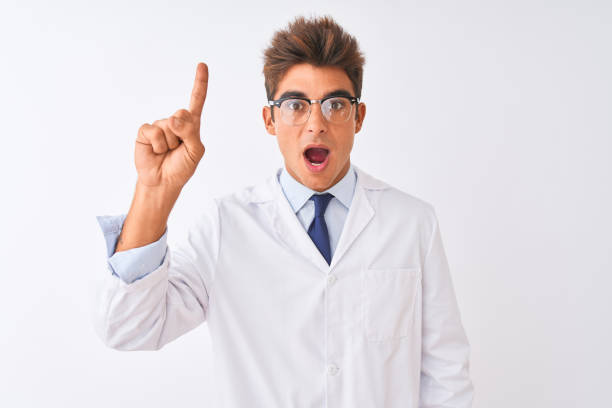 istock Young handsome sciencist man wearing glasses and coat over isolated white background pointing finger up with successful idea. Exited and happy. Number one. 1186061360
