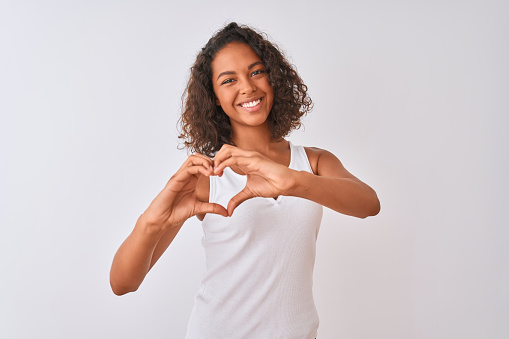 Young brazilian woman wearing casual t-shirt standing over isolated white background smiling in love showing heart symbol and shape with hands. Romantic concept.
