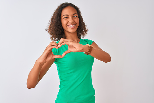Young brazilian woman wearing green t-shirt standing over isolated white background smiling in love showing heart symbol and shape with hands. Romantic concept.