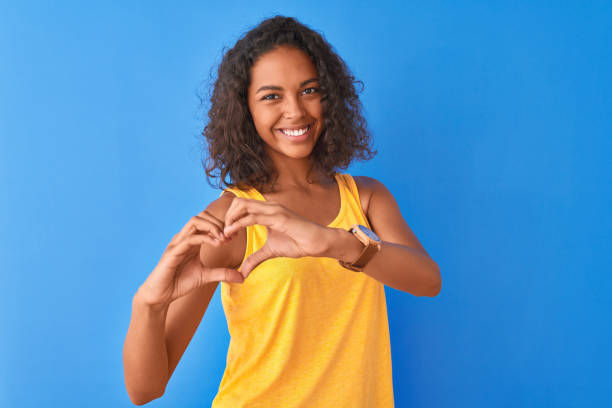 고립 된 파란색 �배경 위에 서있는 노란색 티셔츠를 입은 젊은 브라질 여성이 손으로 심장 기호 모양을하고 사랑에 미소. 로맨틱 한 개념. - teenage girls ideas blue yellow 뉴스 사진 이미지