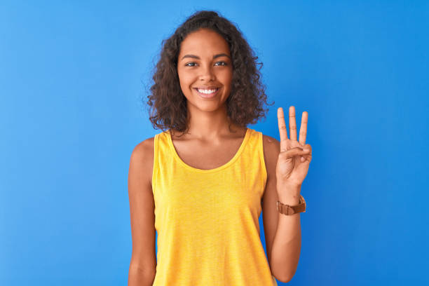 노란 티셔츠를 입은 젊은 브라질 여성이 고립된 파란색 배경 위에 서서 자신감과 행복한 미소를 지으며 손가락 번호 3을 가리키고 있습니다. - teenage girls ideas blue yellow 뉴스 사진 이미지