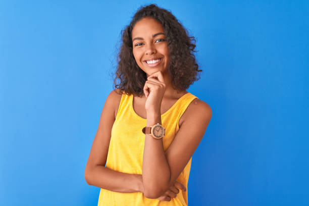 mulher brasileira nova que desgasta o t-shirt amarelo que está sobre o fundo azul isolado que olha confiável na câmera que sorri com os braços cruzados e a mão levantada no queixo. pensando positivo. - teenage girls ideas blue yellow - fotografias e filmes do acervo