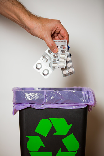 Hand putting expired medicine packages into bin with recycling sign for safe disposal. Person separating dangerous waste. Medical waste management