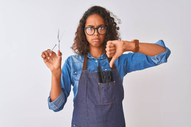 Young brazilian hairdresser woman using scissors standing over isolated white background with angry face, negative sign showing dislike with thumbs down, rejection concept Young brazilian hairdresser woman using scissors standing over isolated white background with angry face, negative sign showing dislike with thumbs down, rejection concept angry hairstylist stock pictures, royalty-free photos & images