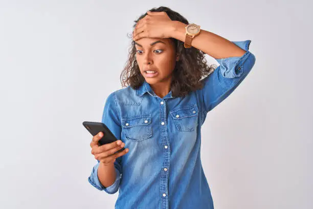 Photo of Young brazilian woman using smartphone standing over isolated white background stressed with hand on head, shocked with shame and surprise face, angry and frustrated. Fear and upset for mistake.